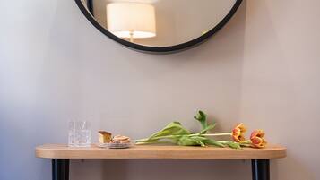 A round mirror on a wall, a small table with a glass and food, and a pink ottoman.