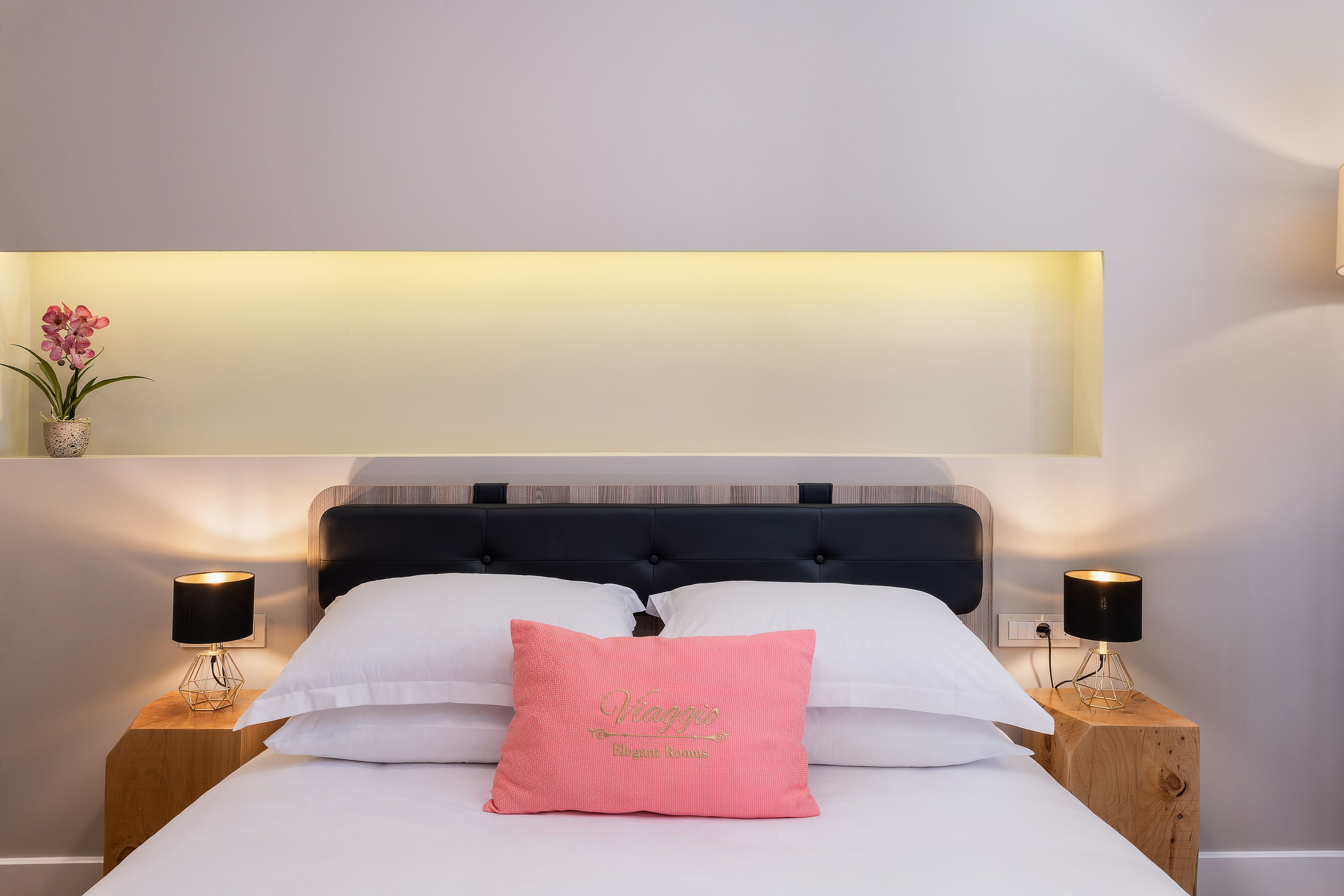 A neatly made bed with white linens, a pink pillow with "Viaggio Elegant Rooms" text, and two bedside lamps on wooden nightstands.