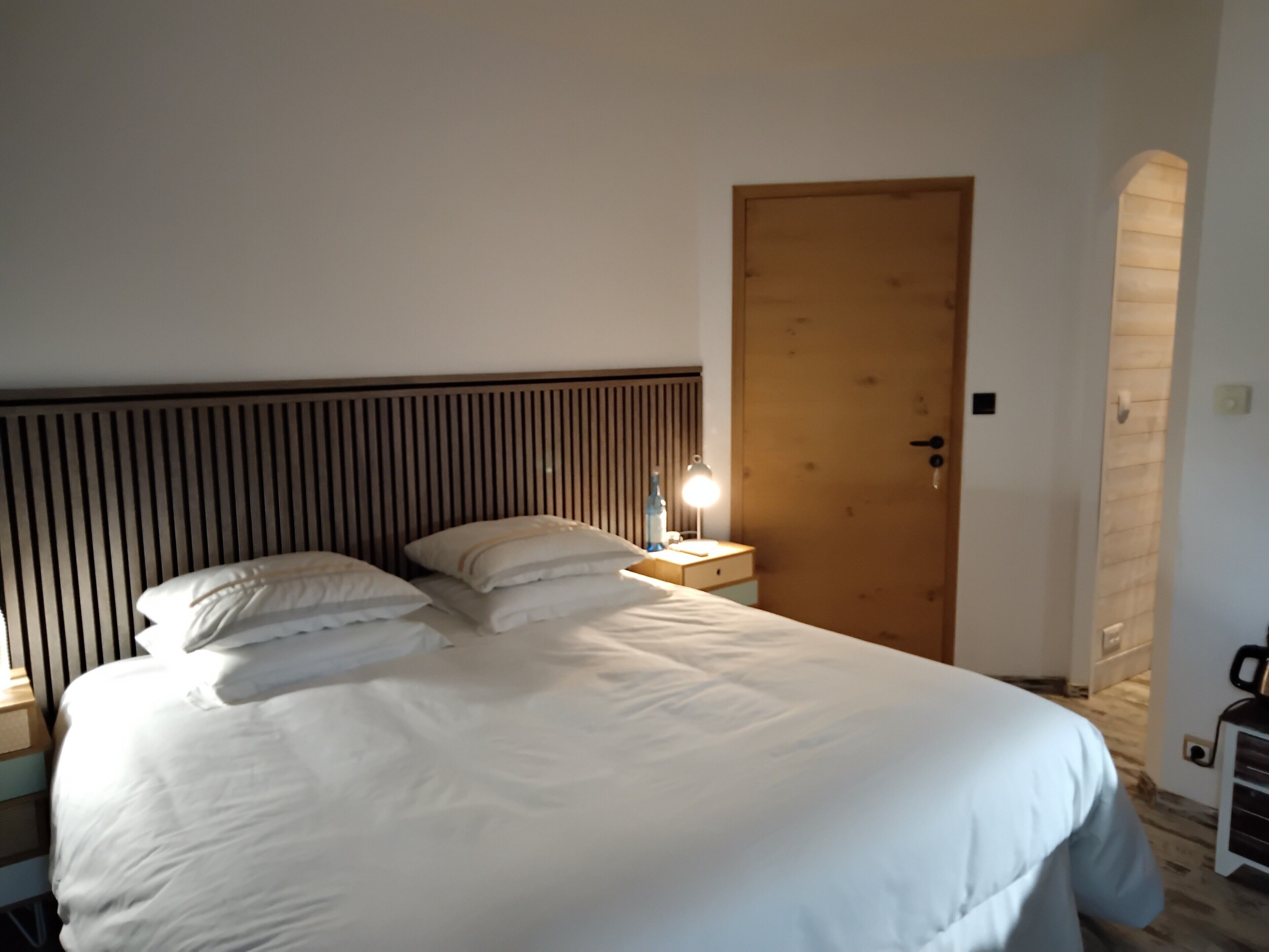 Una cama bien tendida con sábanas y almohadillas blancas, un cabecero de madera, una mesita de noche con una lámpara y una puerta de madera.