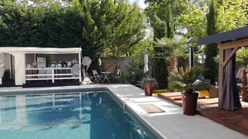 A swimming pool with a covered patio area, outdoor furniture, and a small structure with a table and chairs.
