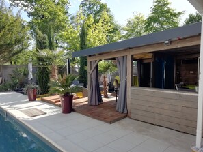 A poolside area with a covered seating area, potted plants, and a wooden deck. - Villa d'Aquitaine Maison d'hôtes (Bretagne-de-Marsan)