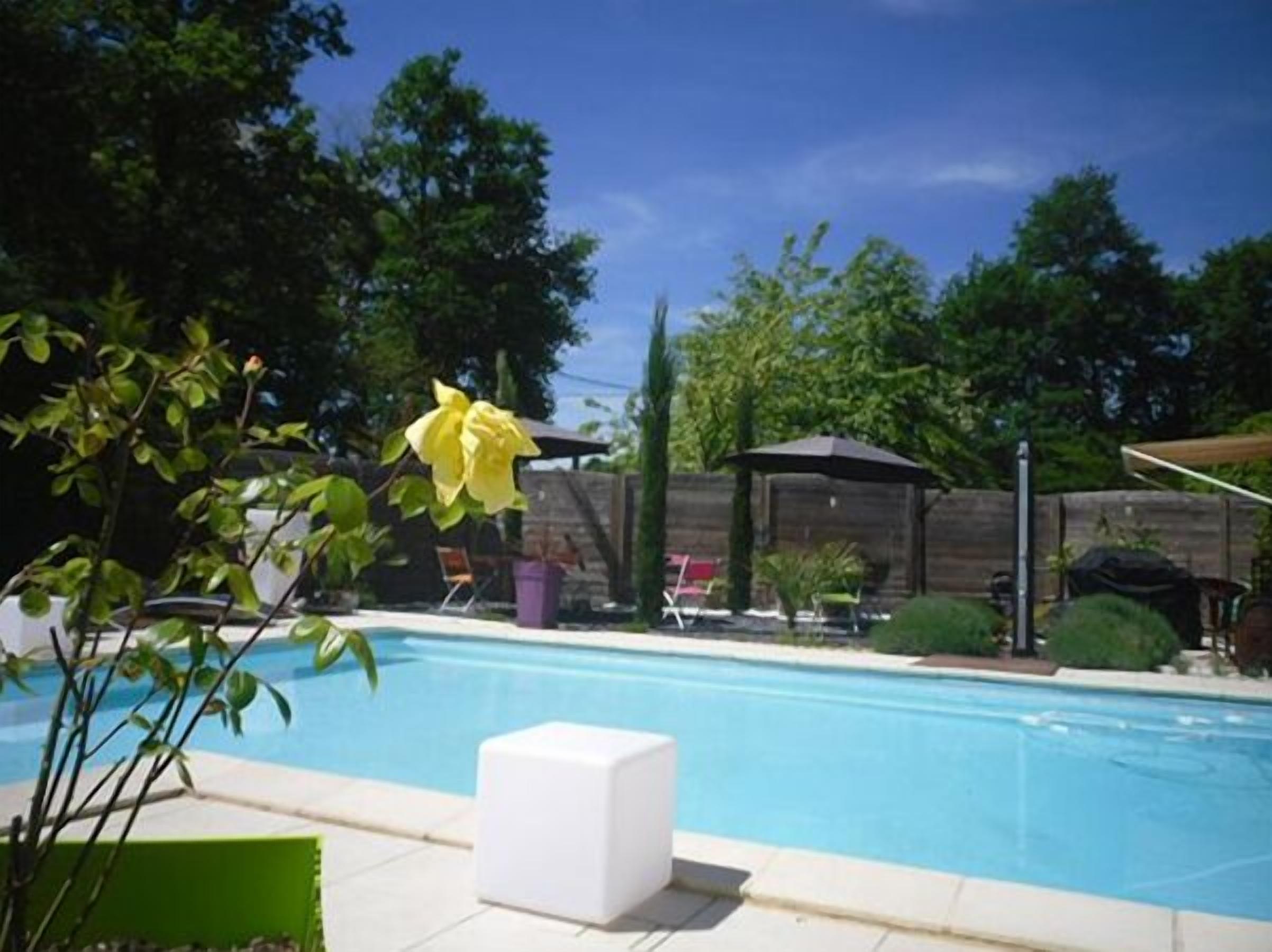 Piscina cercada por vegetação, com uma espreguiçadeira branca e uma área de estar coberta.