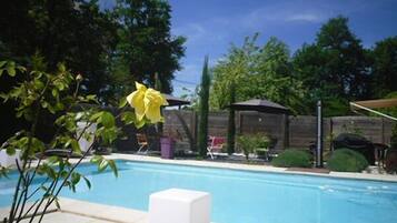 Piscina cercada por vegetação, com uma espreguiçadeira branca e uma área de estar coberta.