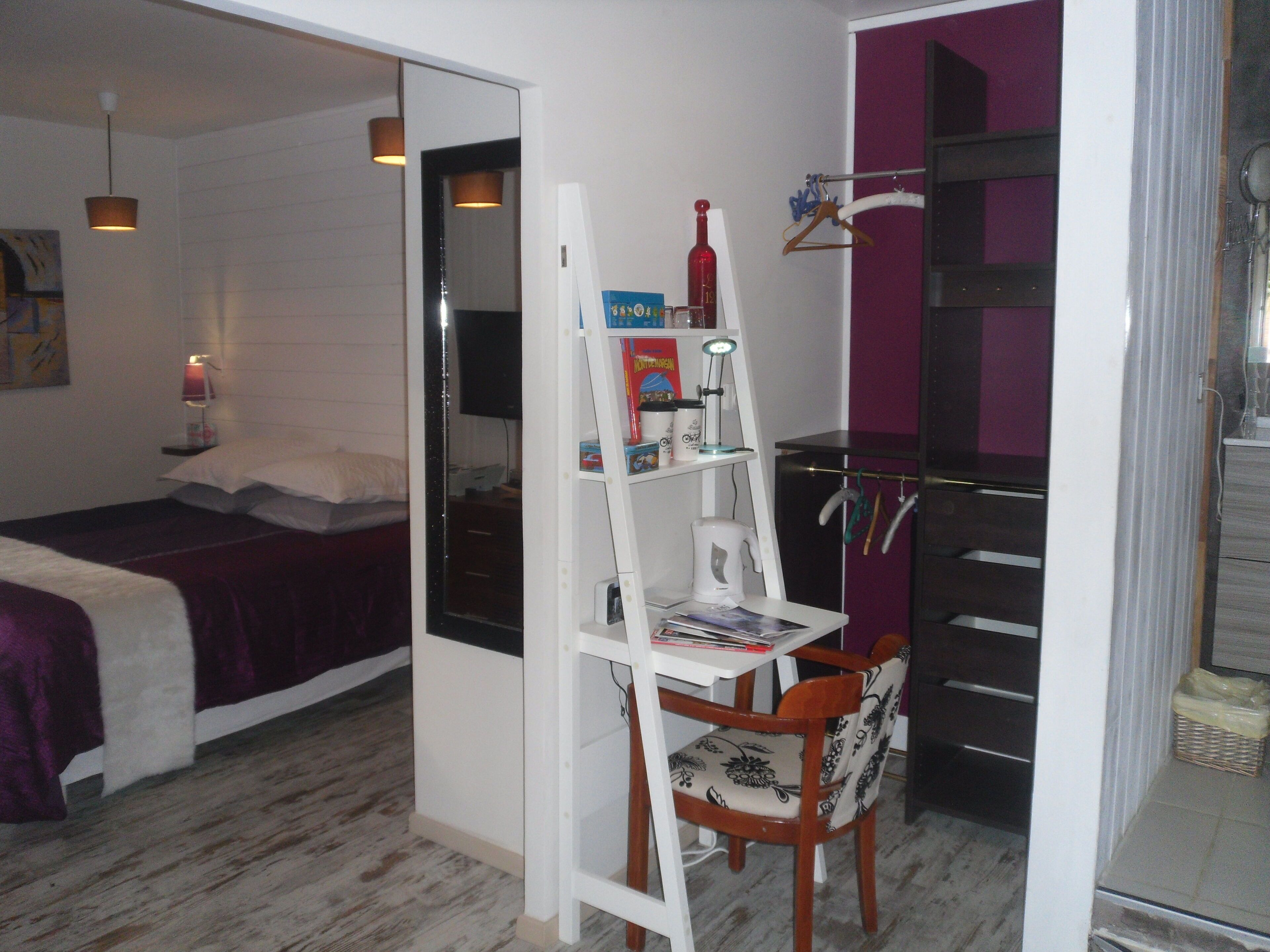 A compact room with a bed, a desk with a chair, a shelf with books and a bottle, and a hanging rack with clothes.