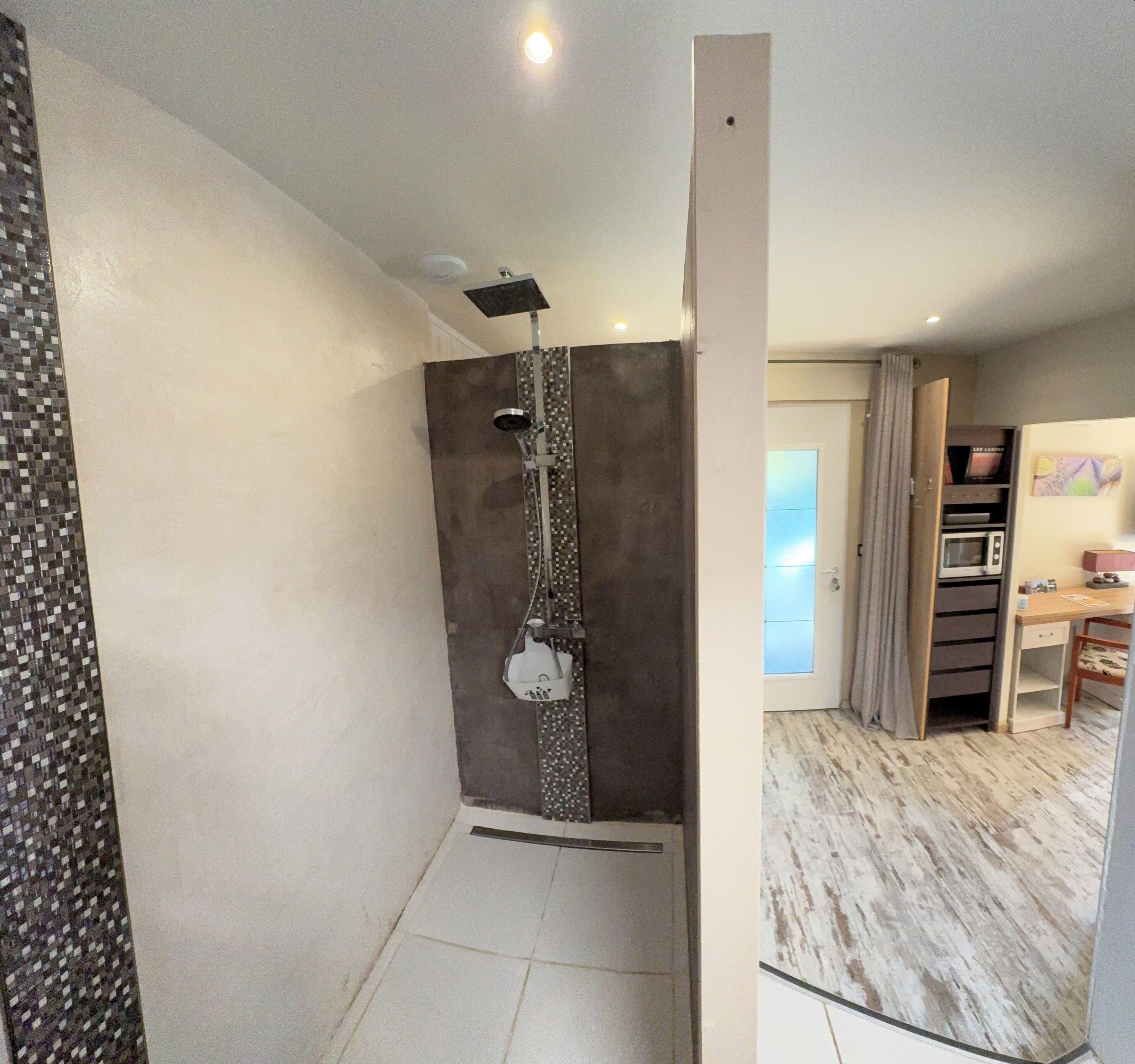 Ein modernes Badezimmer mit Dusche, Toilette und Waschbecken. Ein kleines Regal beherbergt eine Mikrowelle und ein Buch. Der Boden ist mit Fliesen belegt, die Wände sind weiß gestrichen.