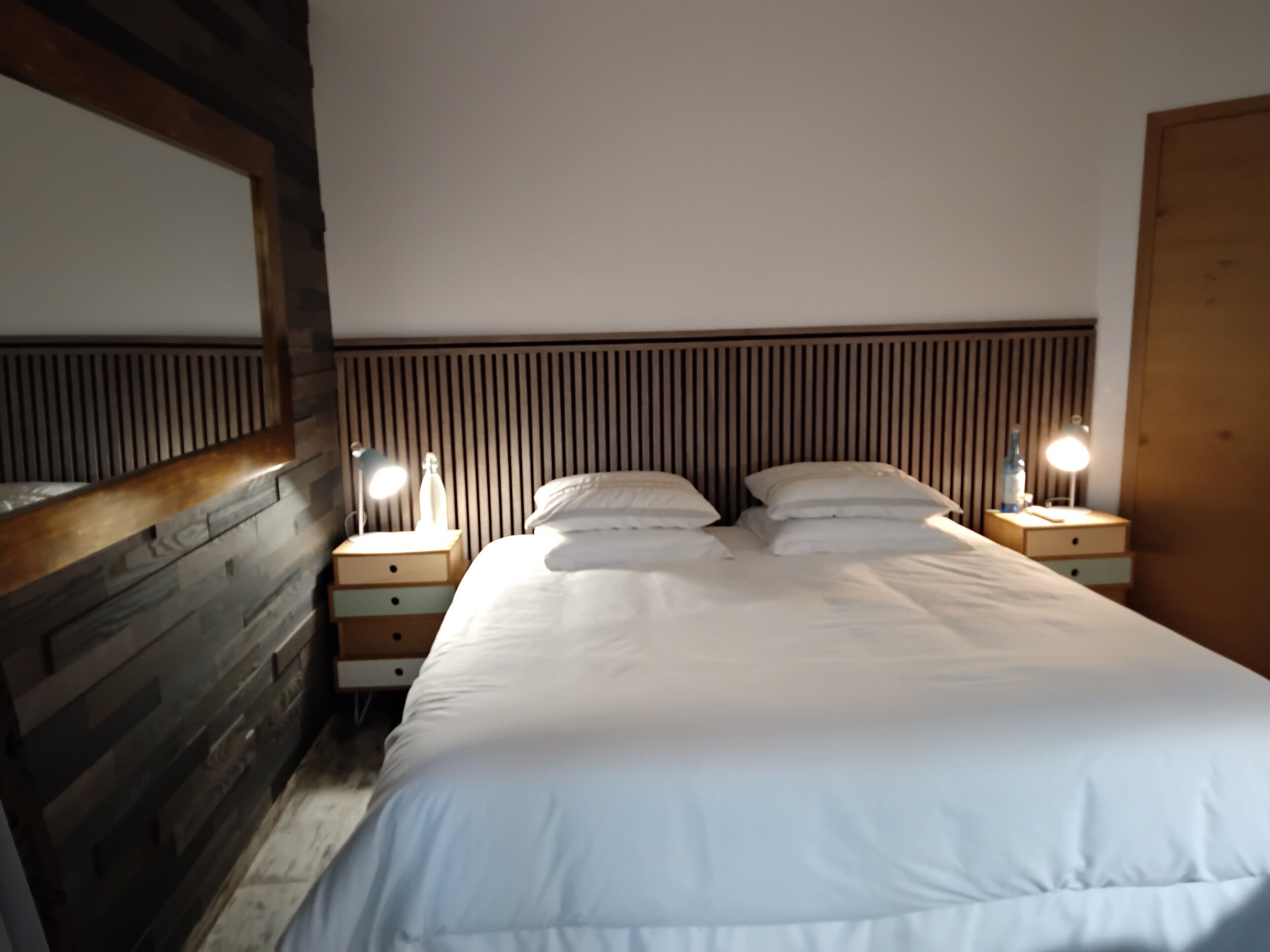 A bedroom with a wooden headboard, a large bed with white bedding, two bedside tables with lamps, and a wooden wardrobe.
