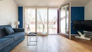 A modern living room with a blue sofa, a glass coffee table, a TV, and a sliding glass door leading to an outdoor area with chairs. - easyhomes - Brera Luxury Suites (Milan)