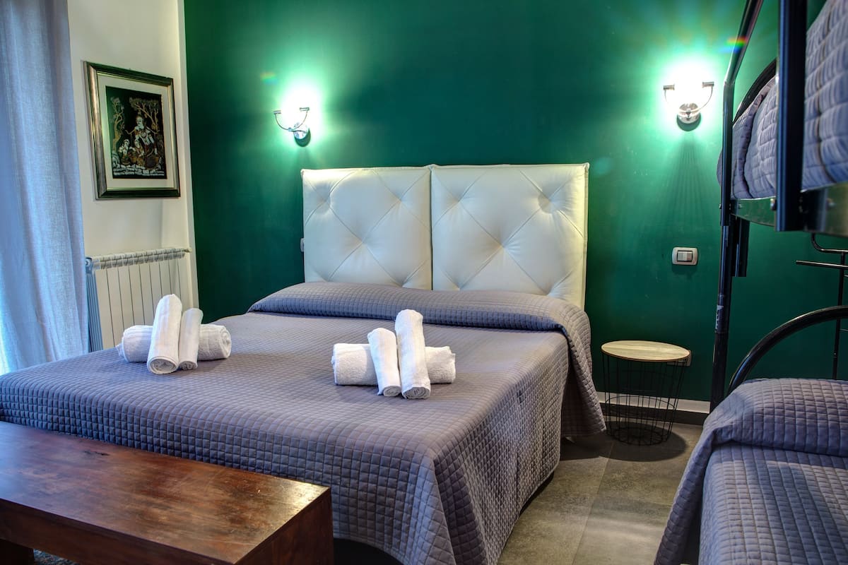 A hotel room with two beds, a wooden bench, a small round table, and a green wall with two wall sconces.
