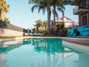 Piscina cristalina con sillas de descanso y palmeras, rodeada por un edificio con balcones.