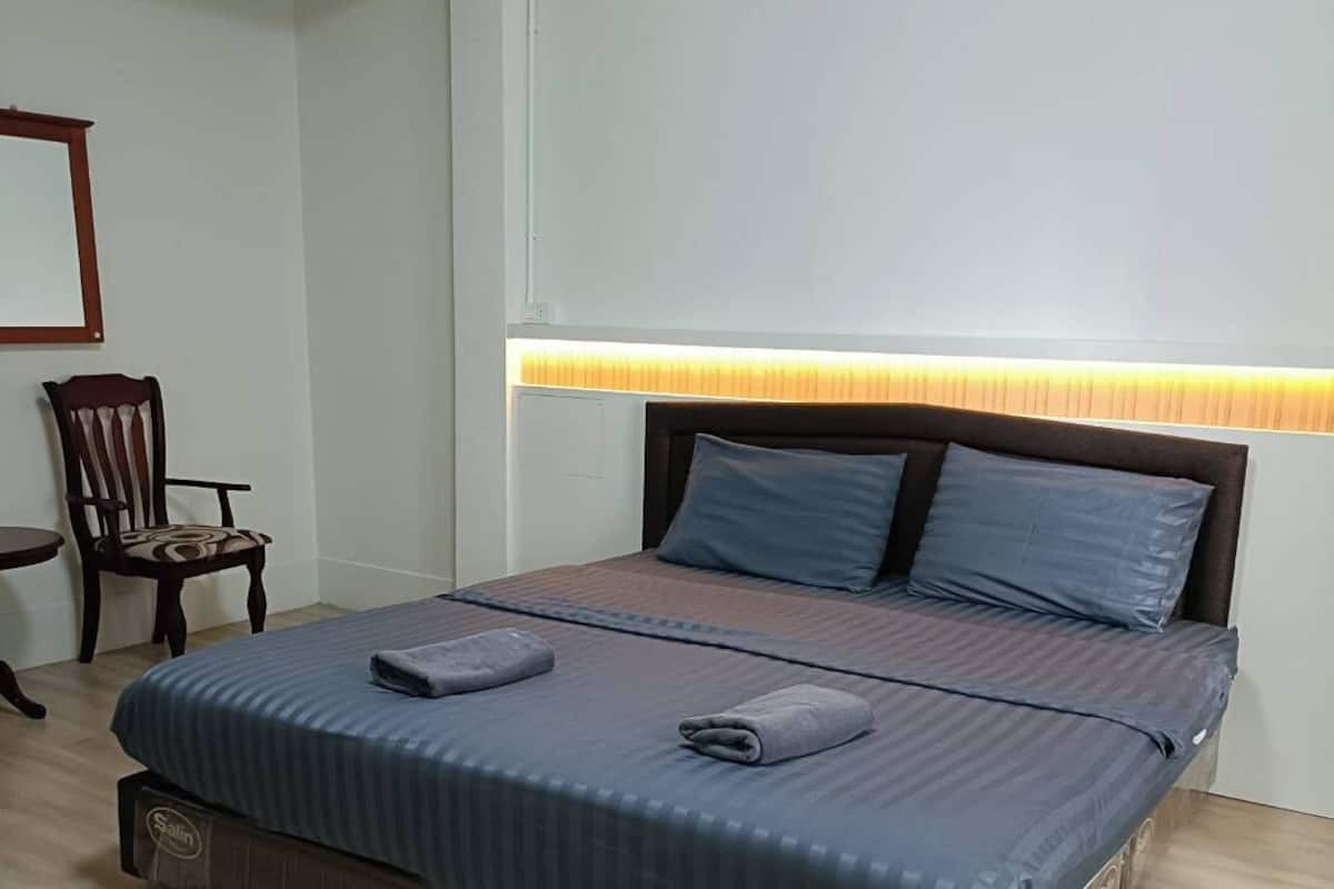 Una cama bien hecha con cabecera, una silla y una mesita de noche en una habitación con paredes y suelo de colores claros.