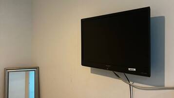 A room with a mounted flat-screen TV on the wall, a bed with a white cover, and a mirror reflecting part of the room.