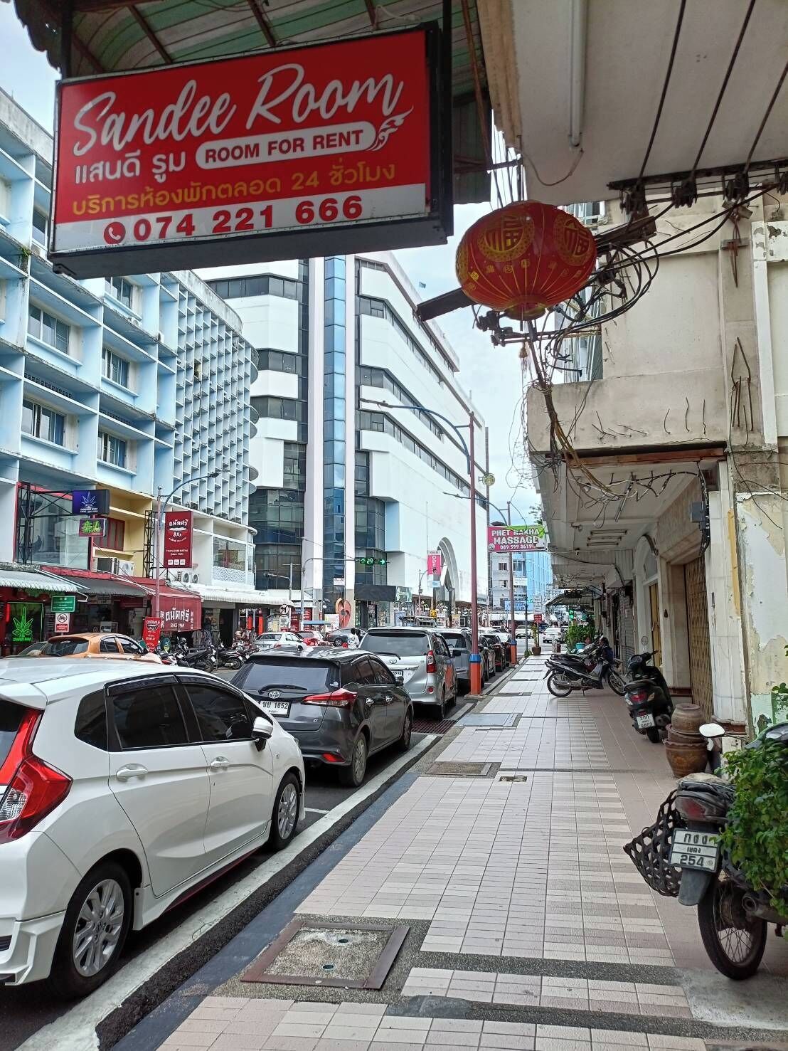 Une vue de rue avec une enseigne indiquant « Sandee Room » à louer, une rangée de voitures garées et des bâtiments ornés de diverses enseignes et publicités.