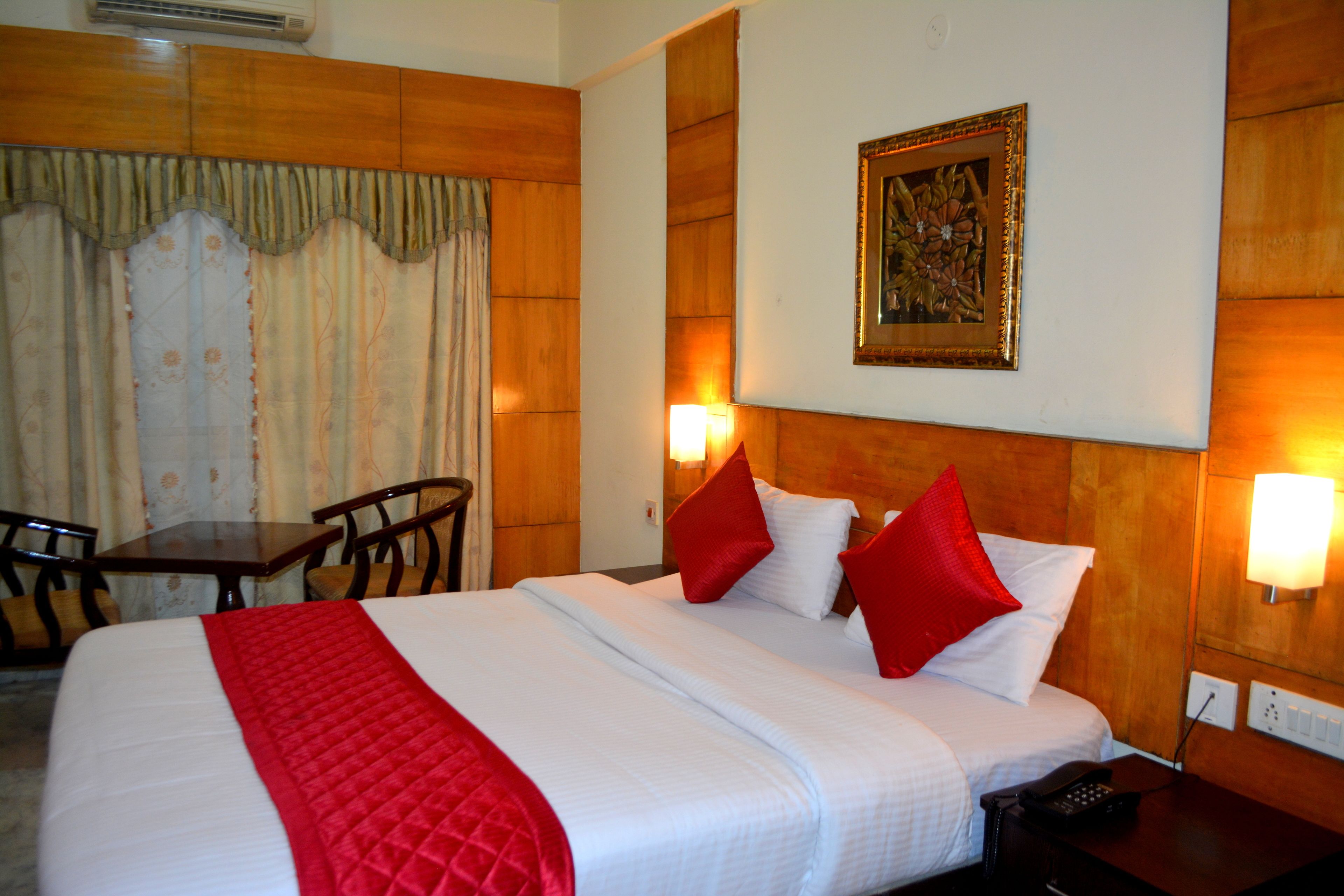 A hotel room with a large bed, two red pillows, a wooden headboard, a painting on the wall, and a desk with chairs.