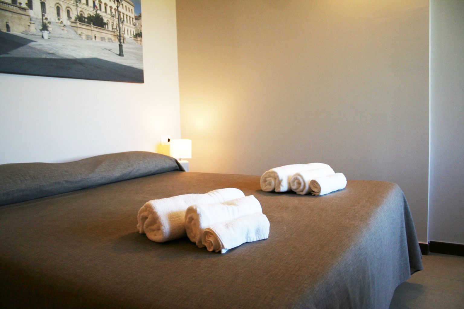 A hotel room with a bed, two rolled towels, a lamp, and a painting on the wall.