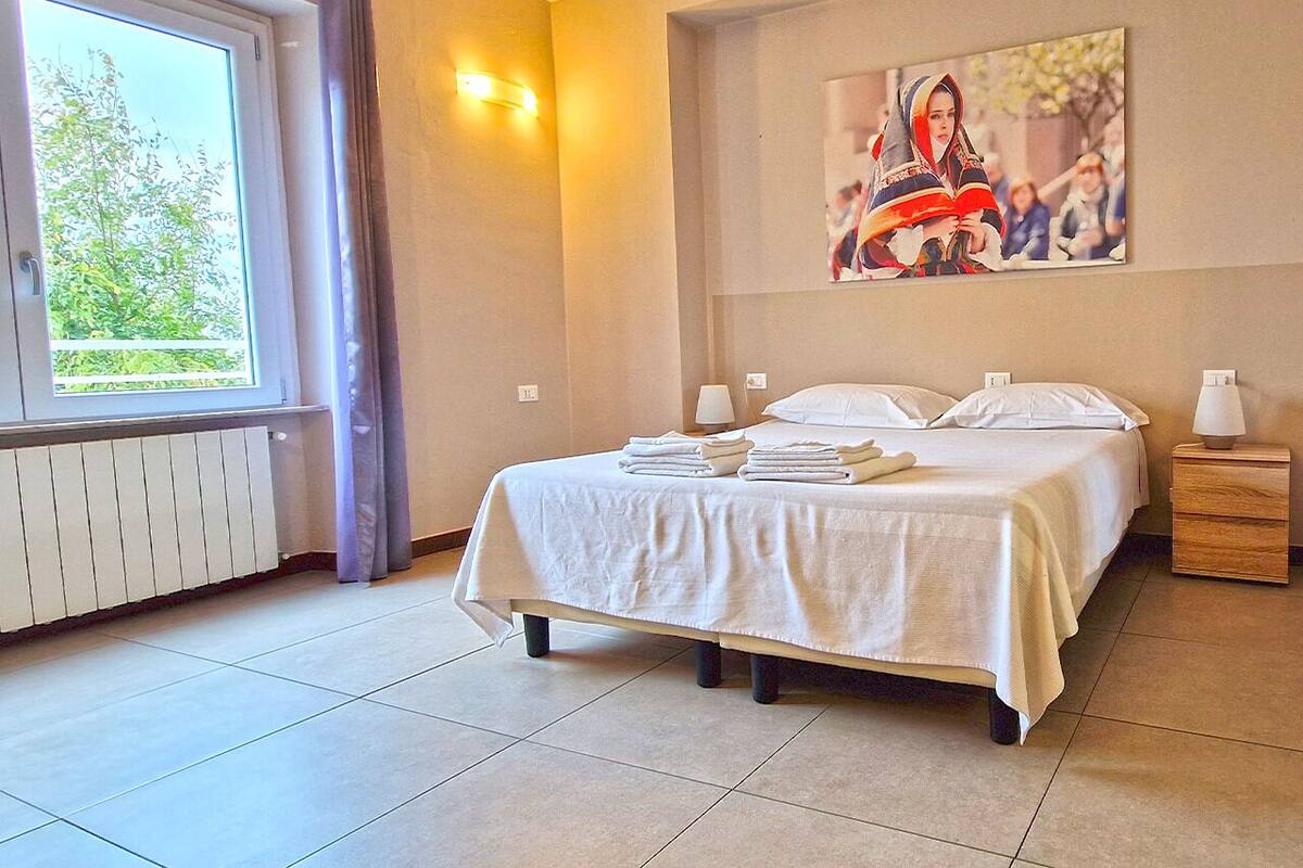 A bedroom with a bed, bedside tables, a window with curtains, a radiator, and a painting on the wall.