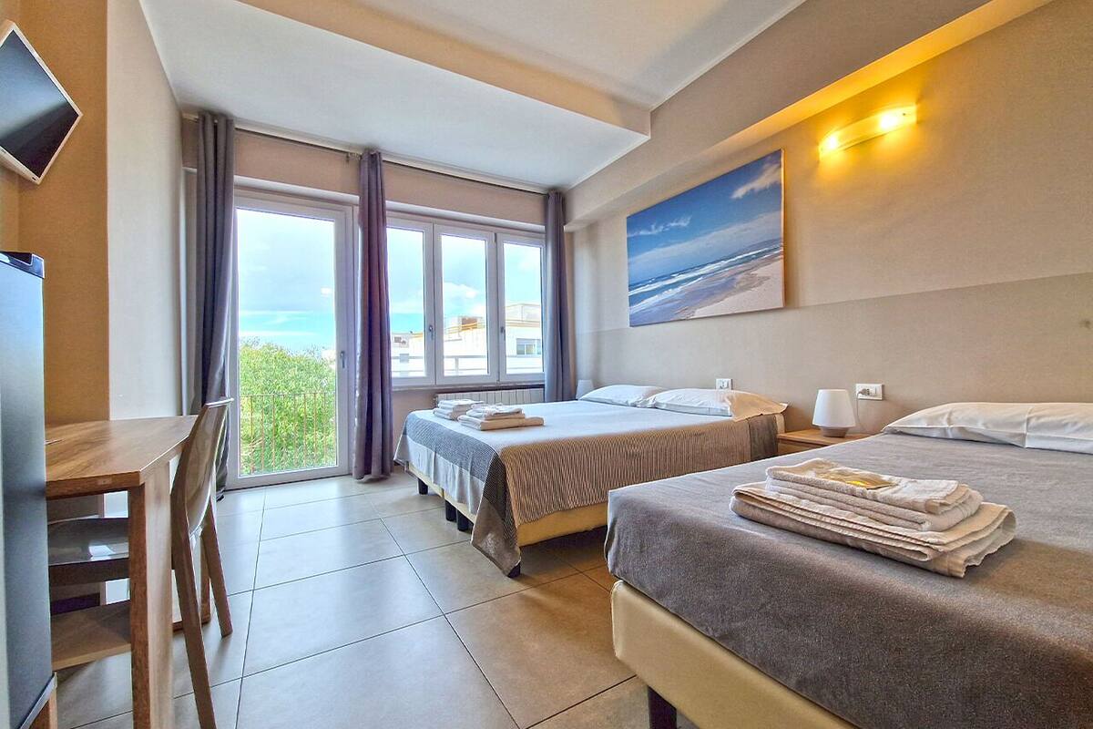A hotel room with two beds, a desk, and a large window with a view of the sea.
