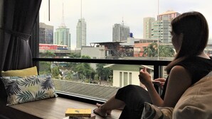 A person sitting by a window with a city view, a sofa with patterned cushions, and a few books on the windowsill. - Cloudy Hostel (Bangkok)