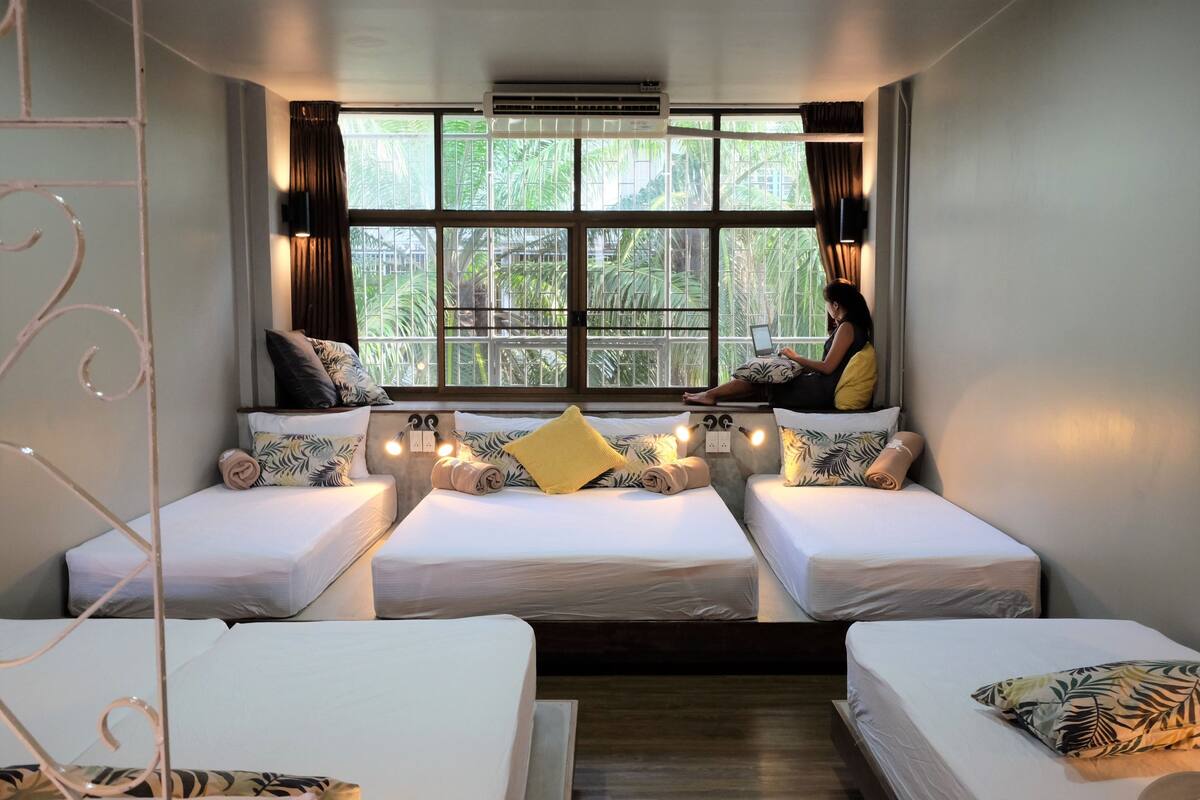 A room with three beds aligned against a window, each with a pillow and a small table.