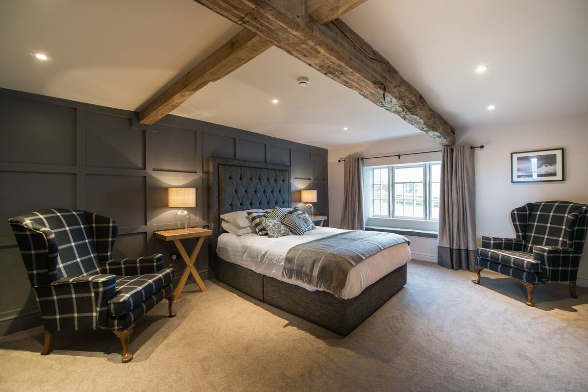 Quarto com uma cama grande, duas poltronas, uma mesa de cabeceira e uma janela com cortinas.