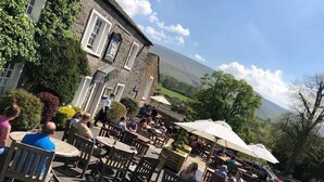 An outdoor cafe with stone walls, wooden tables, and umbrellas, overlooking a scenic landscape. - Assheton Arms (Clitheroe)