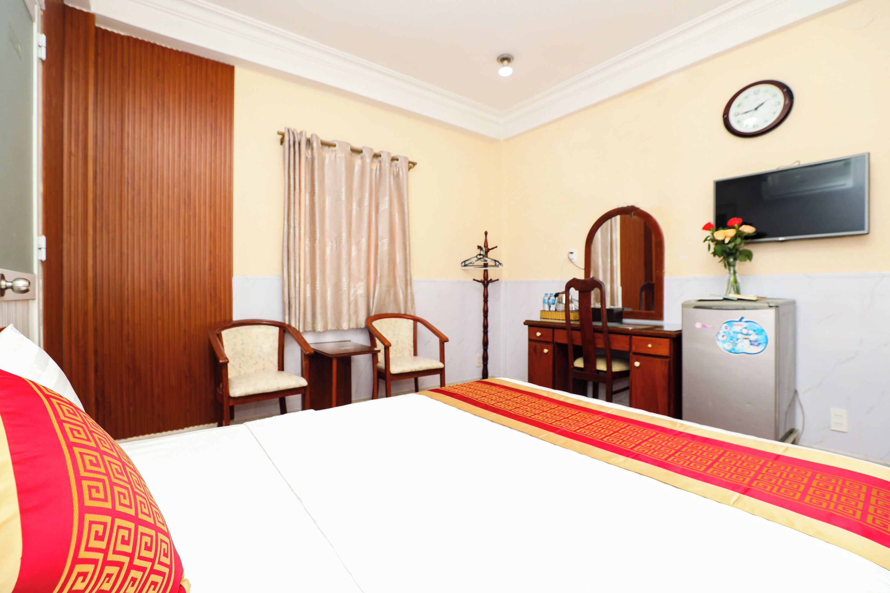 A hotel room with a bed, a desk, a chair, a wardrobe, a television, and a clock on the wall.