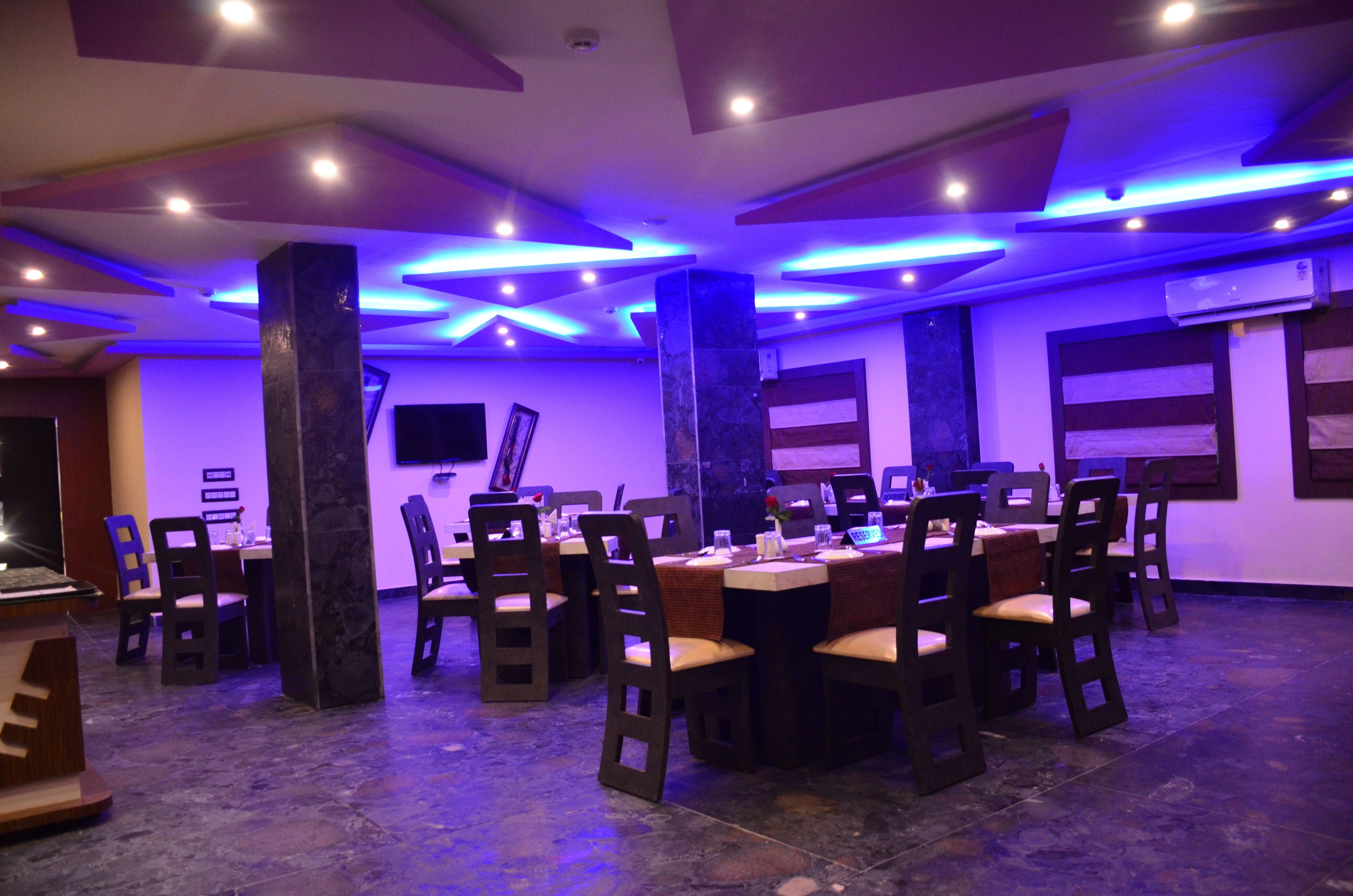 A dining area with round tables and chairs, ceiling lights, and a flat-screen TV on the wall.