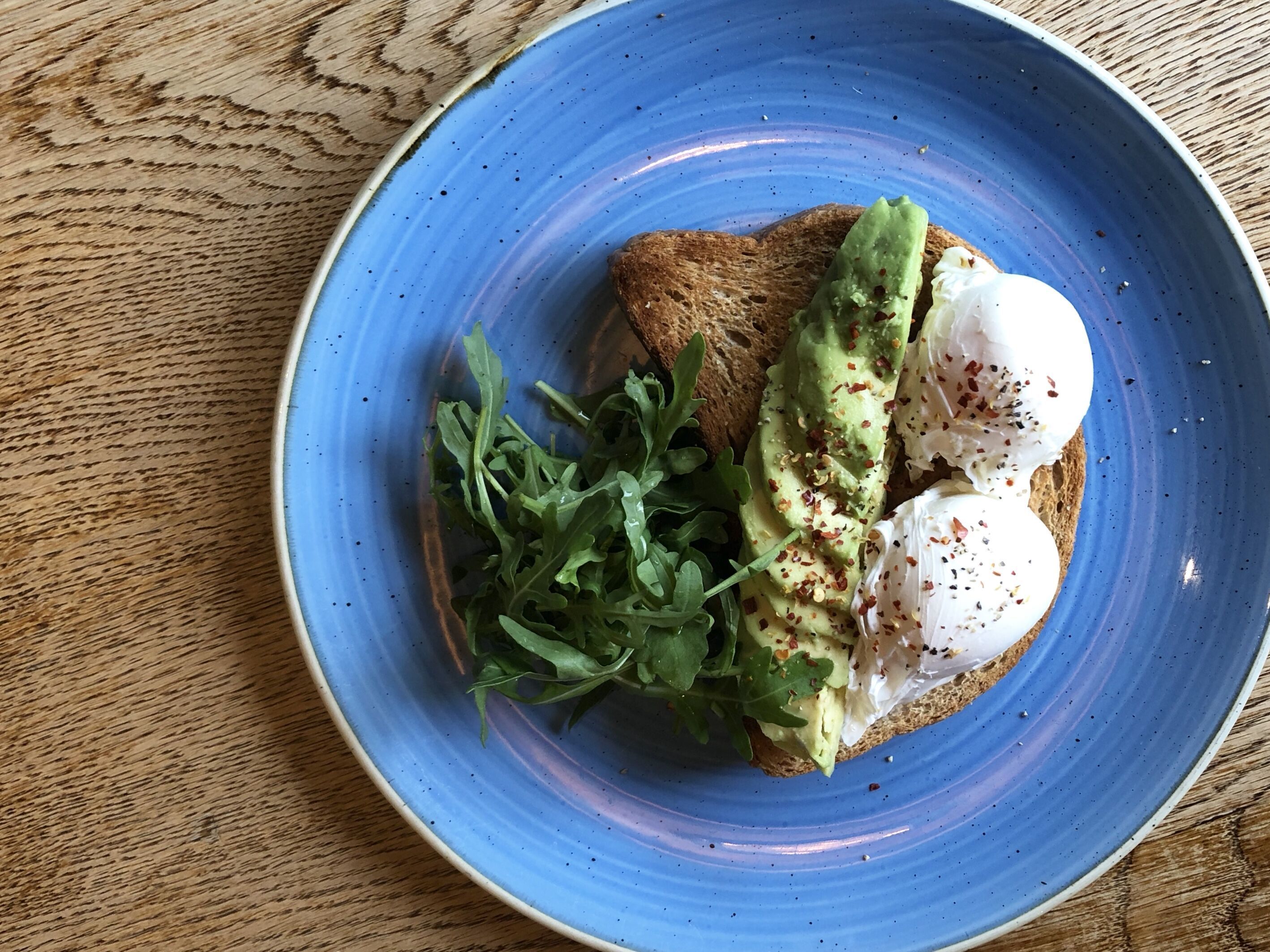 A blue plate with a slice of toast topped with avocado, poached egg, and arugula.