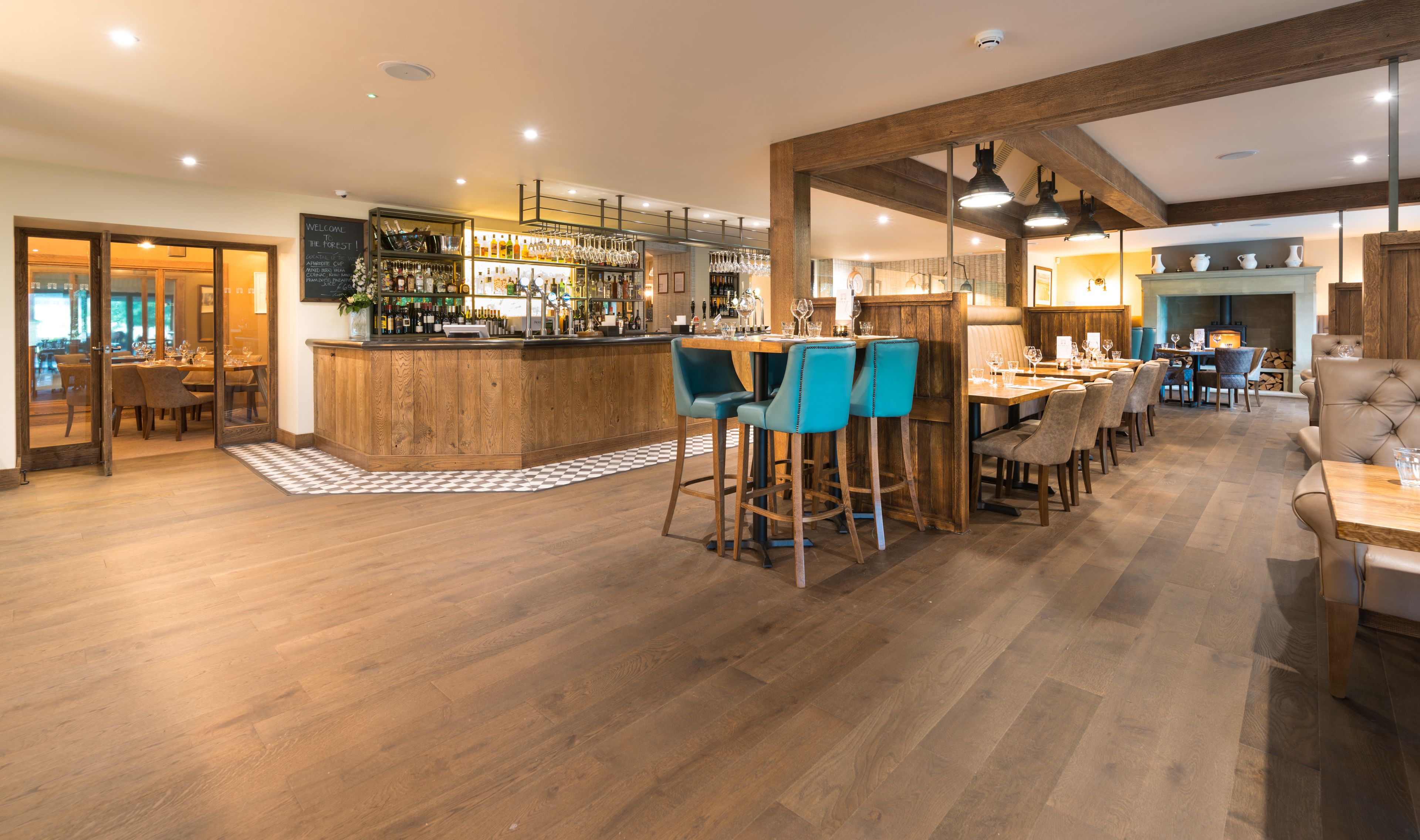 A bar area with wooden flooring, a bar counter, and blue bar stools. There are tables and chairs in the dining area with a checkered rug.