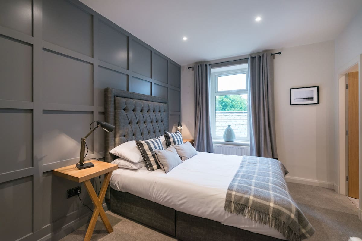 Une chambre avec un grand lit, une table de chevet, une fenêtre avec des rideaux et un tableau accroché au mur.
