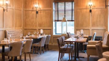 Un espace repas avec des murs lambrés, plusieurs tables entourées de chaises et des luminaires suspendus.