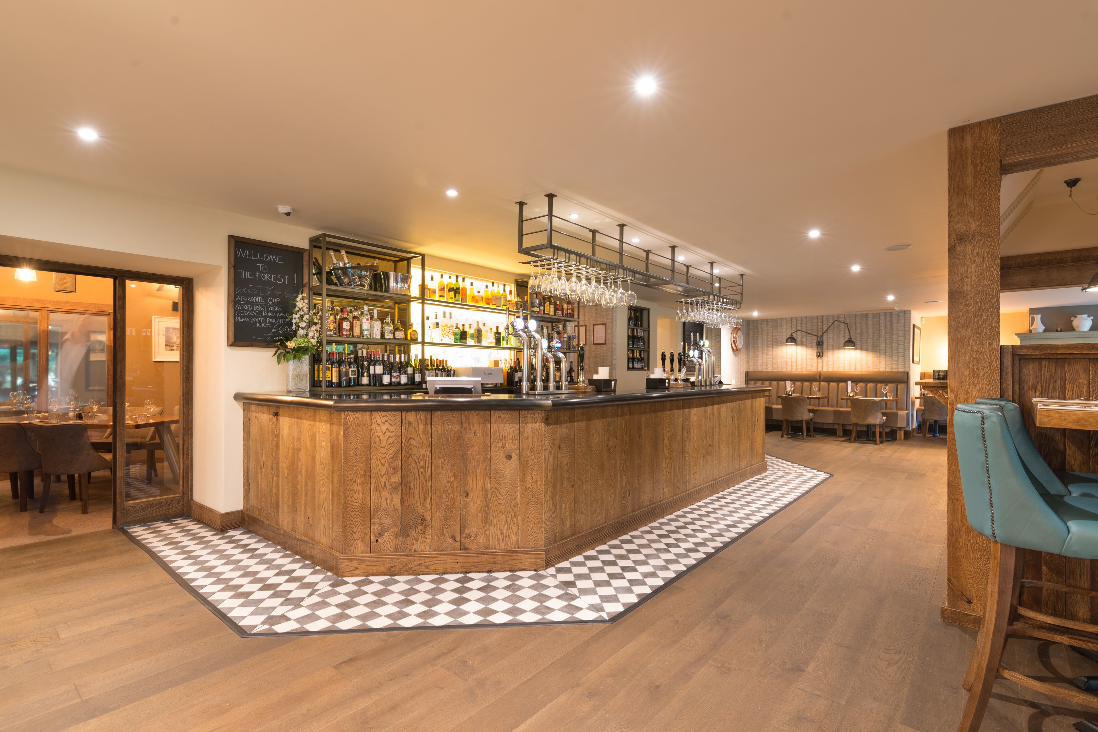 A bar area with a wooden counter, shelves with bottles, and hanging glasses. There is a checkered floor mat in front of the bar.