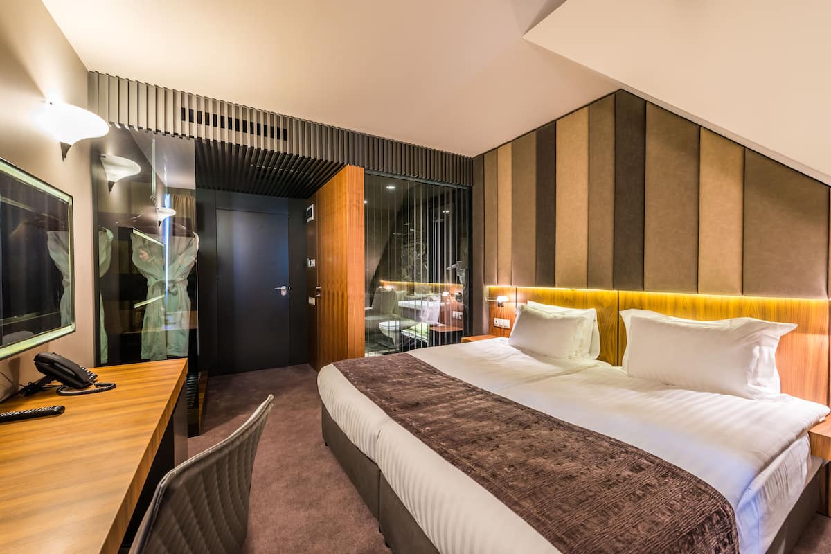 A modern hotel room with a large bed, a desk with a telephone, and a glass partition separating the room from a dining area.