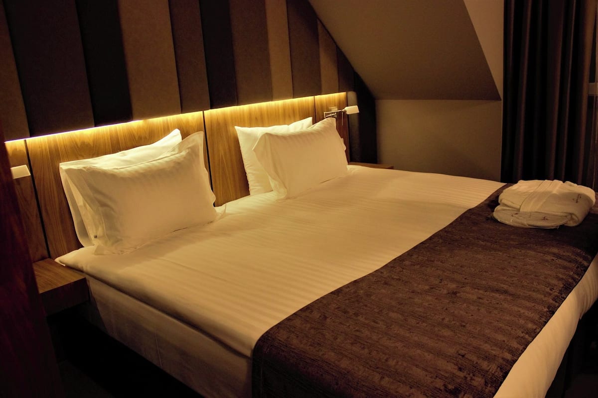 A neatly made bed with white linens and a brown throw blanket, a wooden headboard, and a nightstand with a lamp.