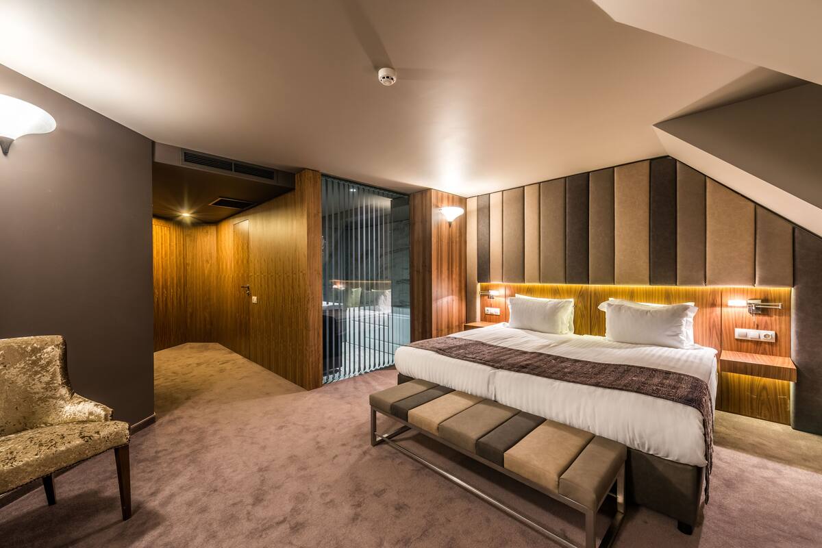 A modern bedroom with a large bed, a bench, a desk, and a view of the city through a glass door.