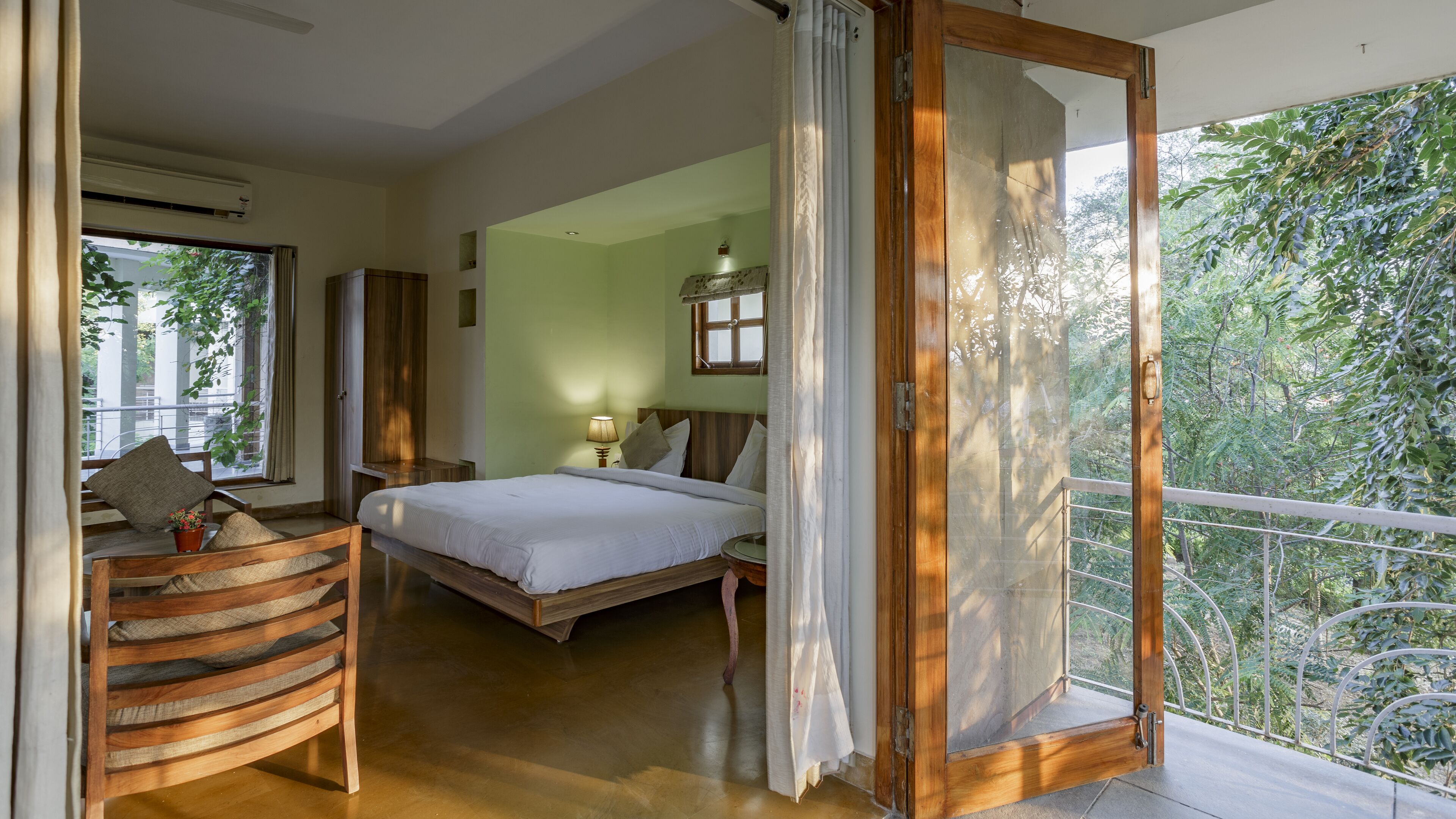 Un dormitorio con cama, una silla de madera, una mesita de noche y una ventana con vista a follaje.