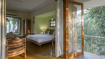 Un dormitorio con cama, una silla de madera, una mesita de noche y una ventana con vista a follaje.