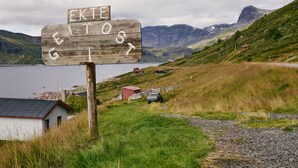 Een houten bord met de tekst 'GEITOST', gelegen in een landelijke omgeving met een meer, bergen en enkele gebouwen op de achtergrond.