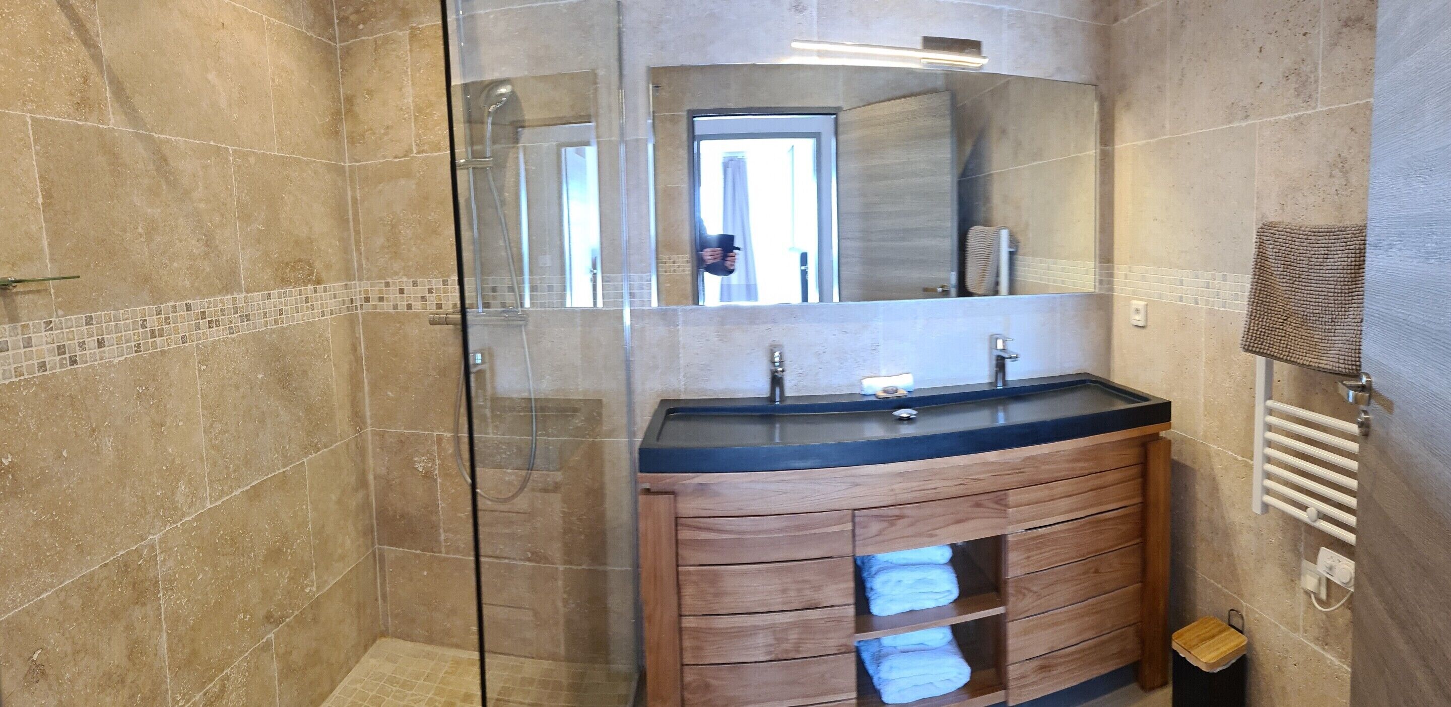 Un baño moderno con ducha envuelta en vidrio, tocador de madera con encimera negra y un perchero en la pared.