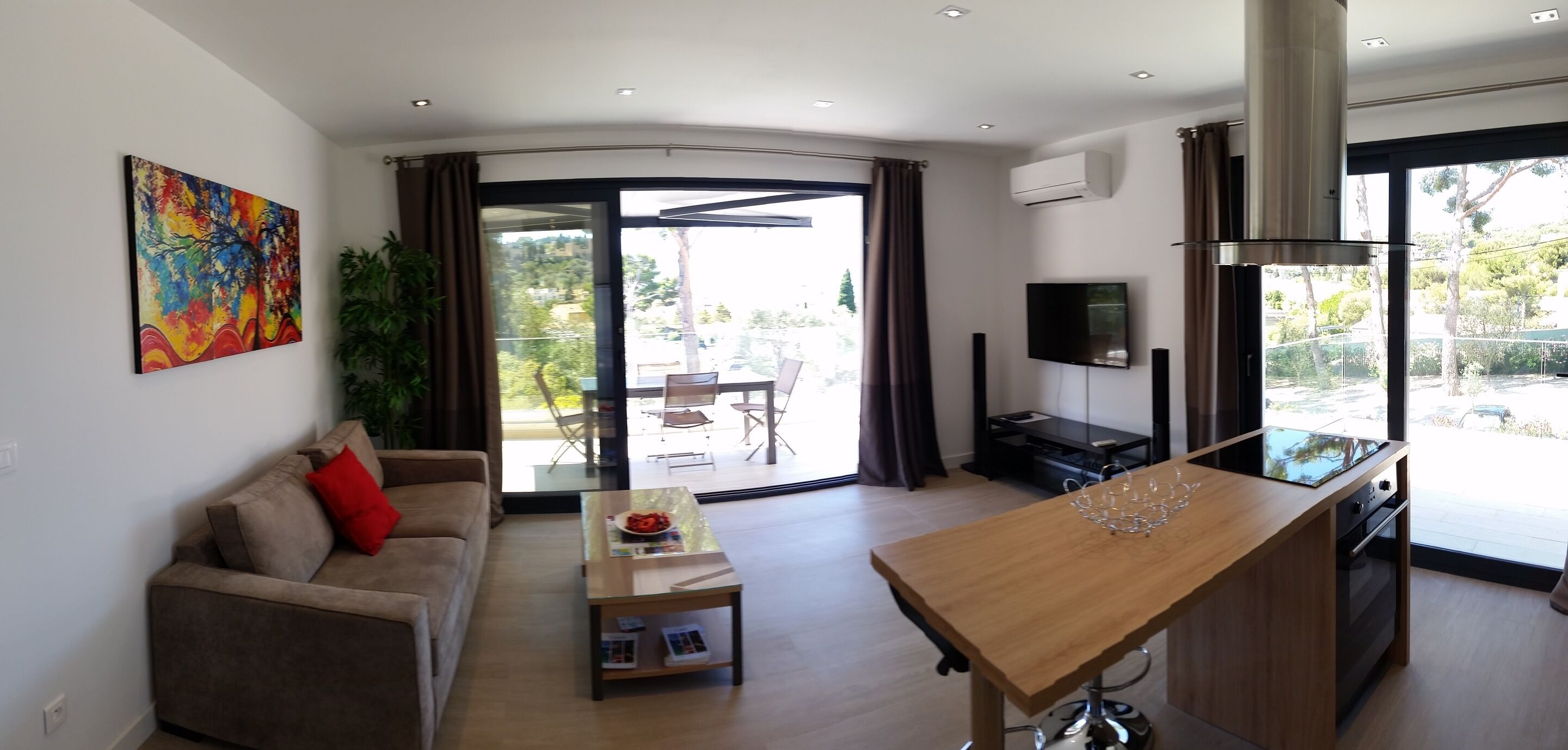 A modern living room with a sofa, coffee table, and a dining area with a wooden table. There is a TV mounted on the wall and a large window providing a view of the outdoors.