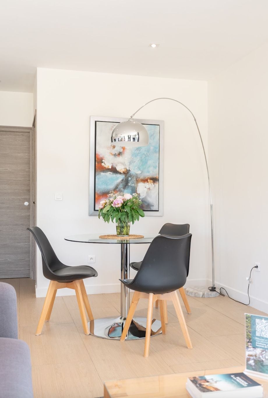 Un espace repas moderne avec une table en verre, deux chaises noires, un vase de fleurs et un lampadaire.