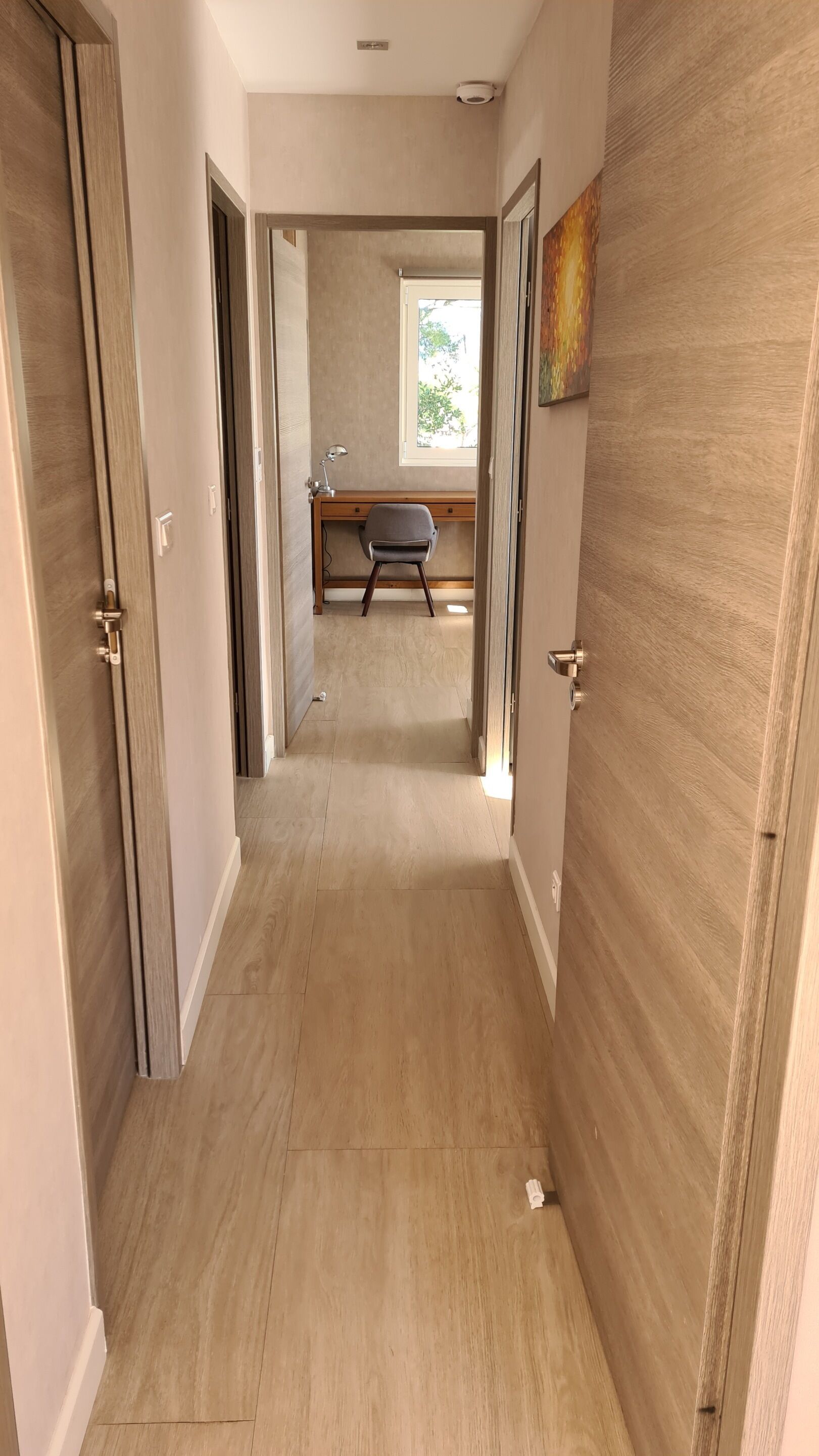 Corredor com soalho de madeira e portas de ambos os lados, dando acesso a um quarto com escrivaninha e cadeira.