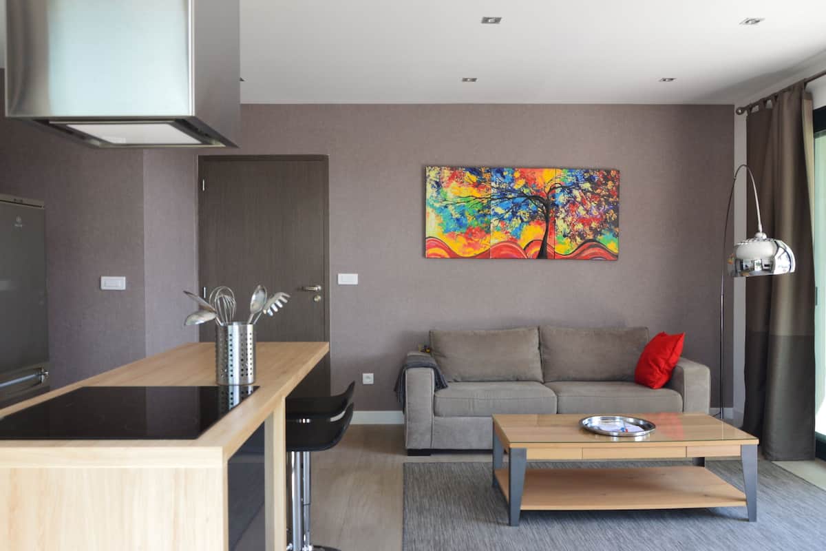 A modern living room with a sofa, a coffee table, a painting, and a kitchen area with a stove.