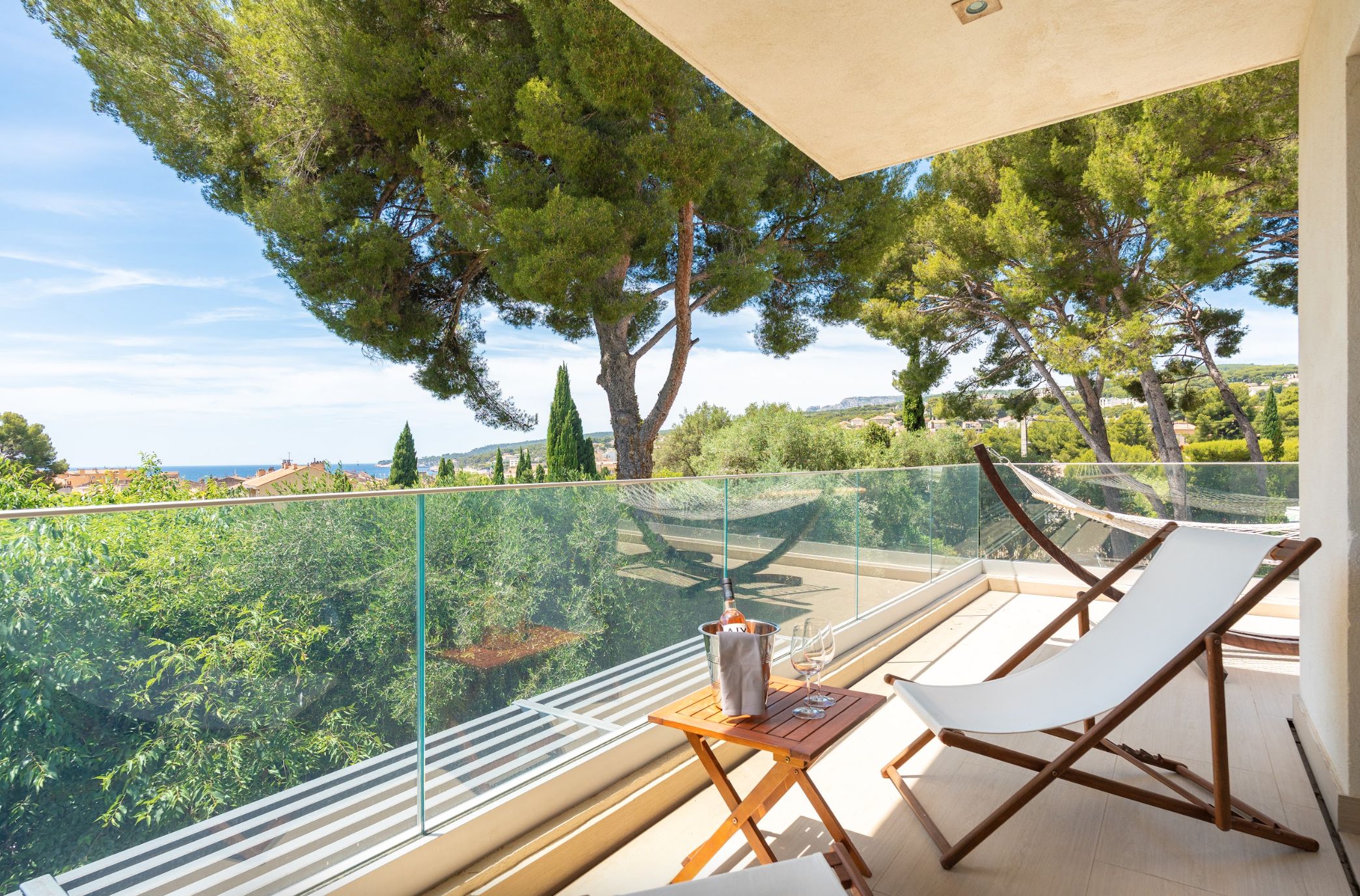Een balkon met een glazen hekwerk, twee witte lounge fauteuils, een houten tafel met een fles en glas, en uitzicht op een groen landschap met bomen en gebouwen.