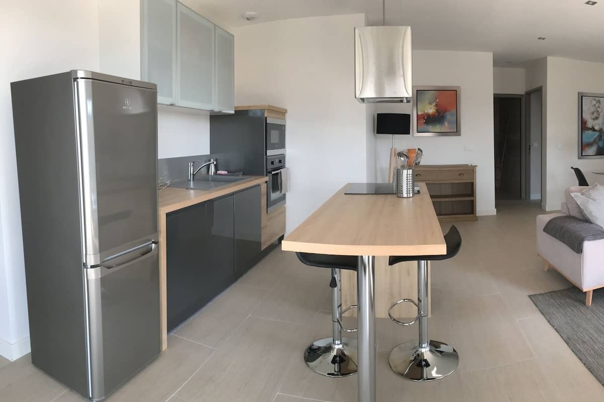 A modern kitchen with a refrigerator, stove, and bar stools. There is a dining area with a table and chairs, and a living room with a sofa and coffee table.