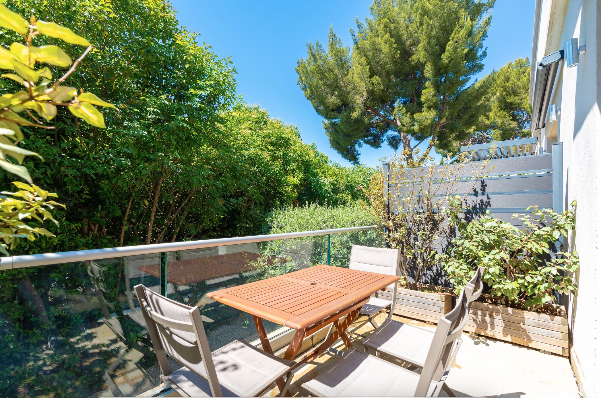 An outdoor patio with a wooden table and chairs, surrounded by greenery and a clear sky.