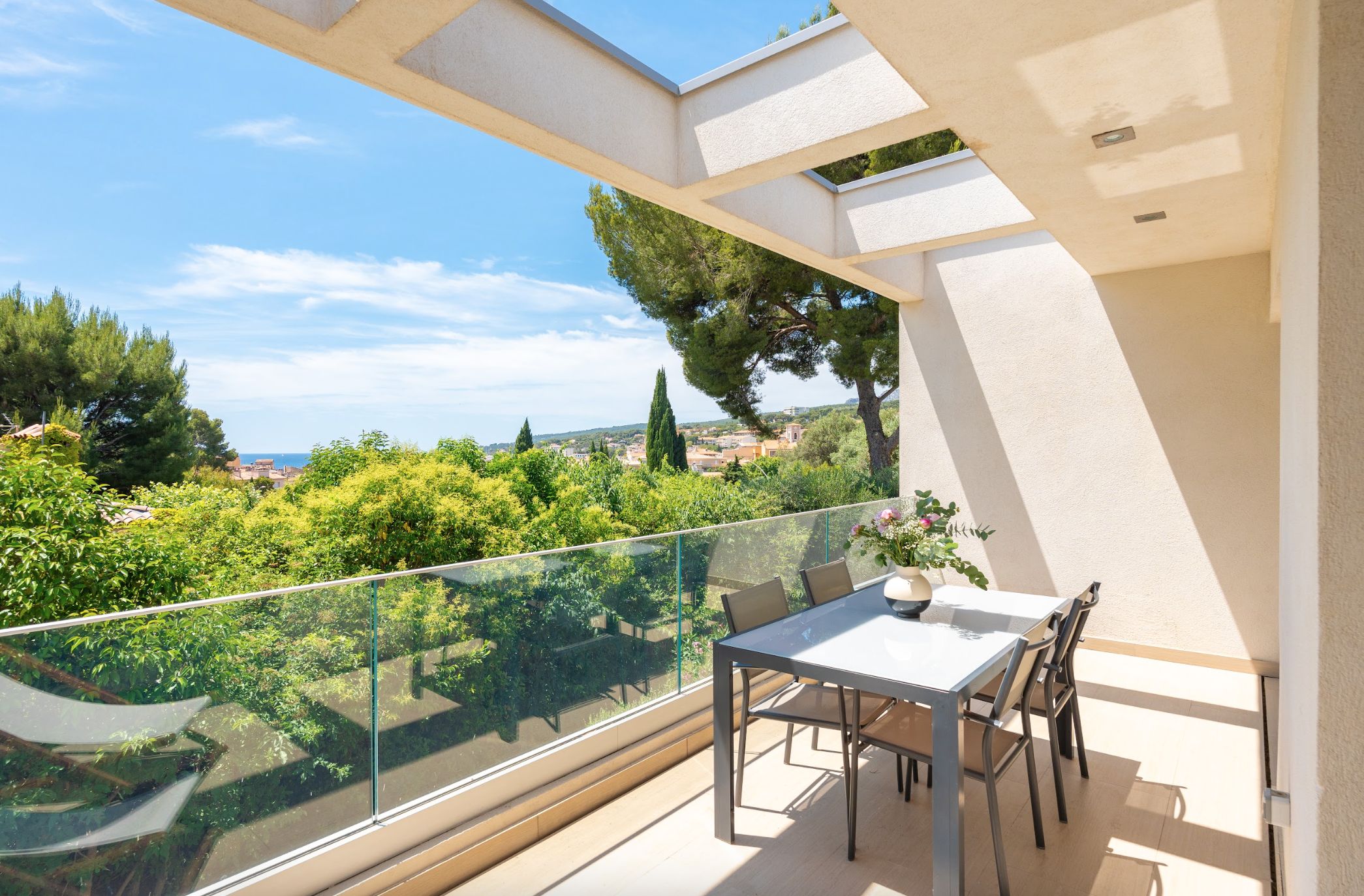 En balkon med gelænder af glas, et bord og stole, med udsigt over et frodigt grønt landskab og en by i det fjerne.