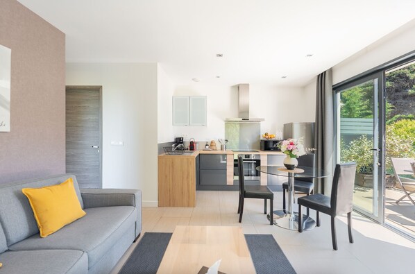 A modern kitchen with a dining area, a grey sofa with a yellow pillow, and a large window offering a view of greenery.