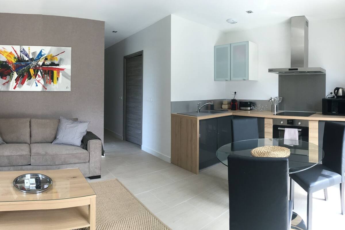 A modern living room with a sofa, coffee table, and a colorful abstract painting. A kitchen area with a microwave, stove, and cabinets is visible.