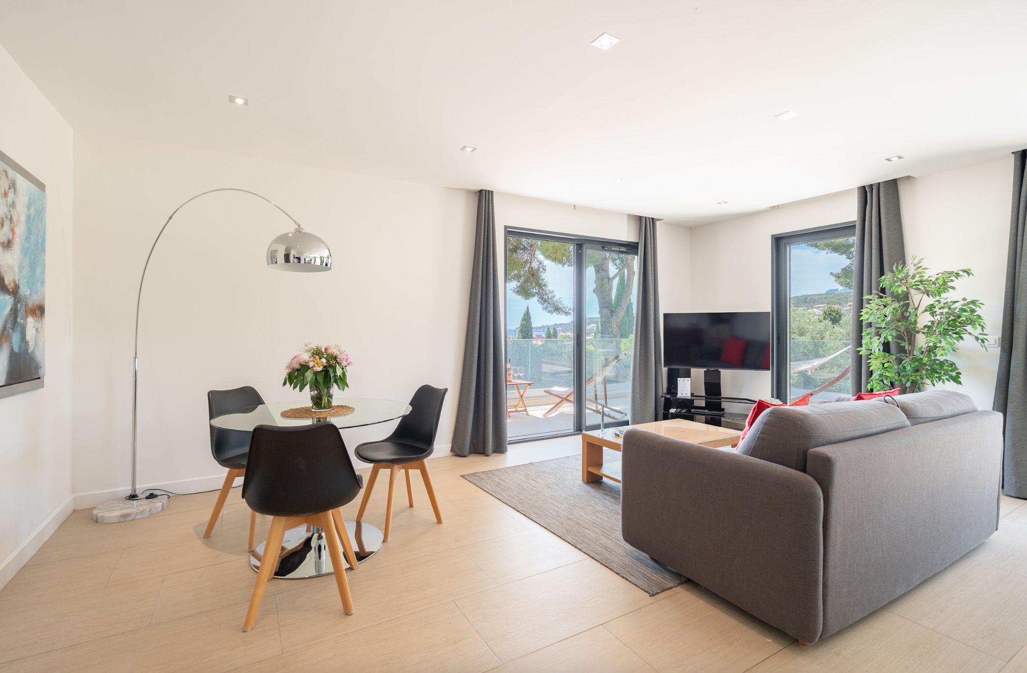 A modern living room with a grey sofa, a dining table with chairs, a floor lamp, and a TV.