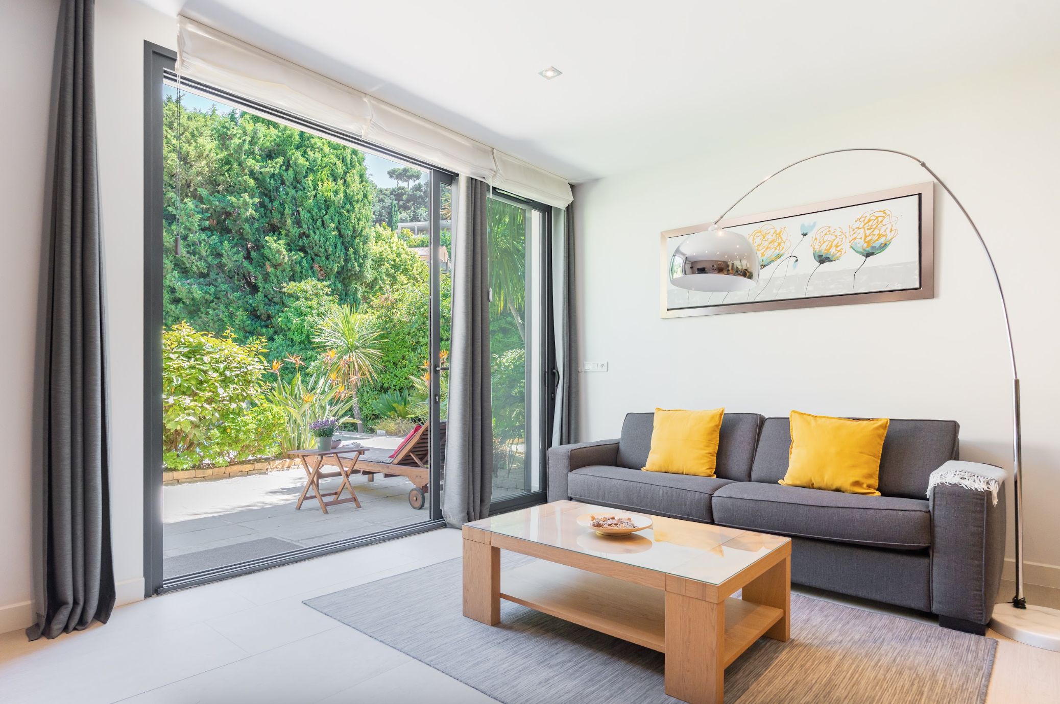 Una sala moderna con un sofá gris, cojines amarillos, una mesa de centro de vidrio y vistas a un jardín a través de grandes puertas correderas.